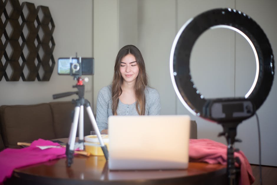 A young woman vlogging indoors using a smartphone, tripod, and ring light setup.